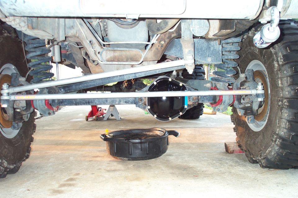 Under-front view of lifted 4x4 solid axle with black diff cover, steering linkage, and drain pan below