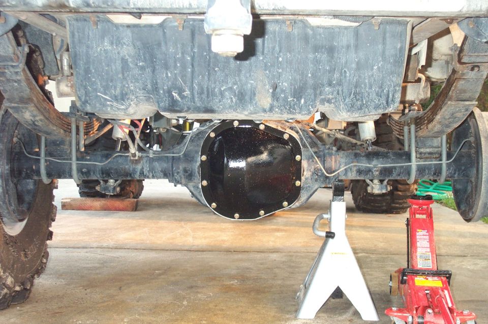 Underside rear view of a lifted truck's solid axle with fresh black differential cover, supported near a jack stand