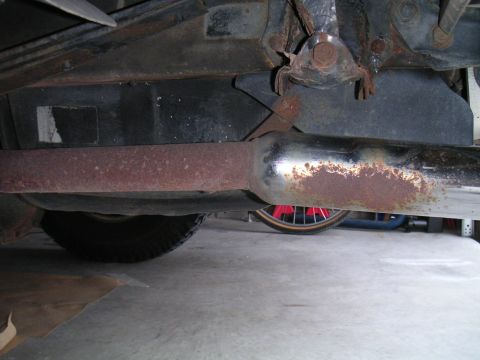 1994 Ford Bronco - Underbody view looking along a heavily surface-rusted exhaust pipe and muffler running beneath a vehicle's rocker and floor pan on a concre…