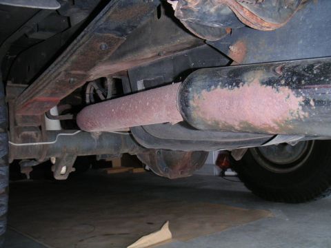 1994 Ford Bronco - Low underbody view of a rusty muffler and tailpipe running alongside the frame rail and leaf spring of a truck parked on a concrete garage…