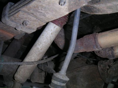 1994 Ford Bronco - Underside close-up of a white shock absorber with rust speckling, mounted through a rusty frame bracket next to a sway bar end link with a…