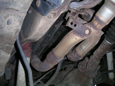 1994 Ford Bronco - Underbody shot looking up at a rusty dual exhaust downpipe assembly with a bolted hanger bracket and flange, routed alongside the frame rai…