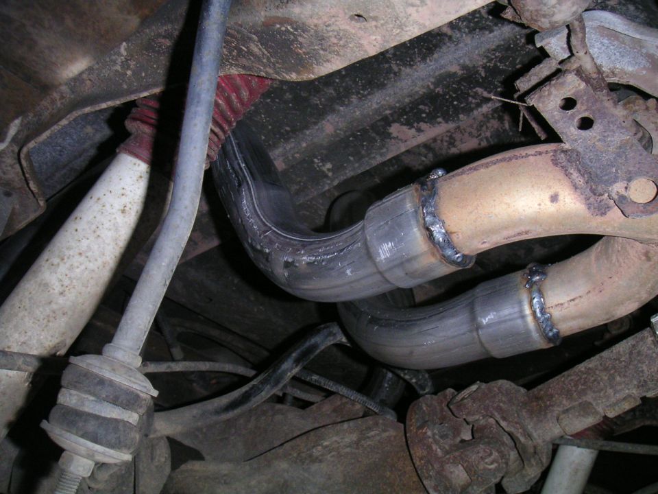 Freshly welded dual exhaust pipes under a truck beside a white shock and sway bar