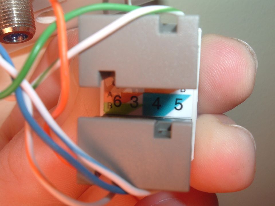 Hand holding RJ45 keystone jack showing T568A/B color-code pin map and twisted-pair wires