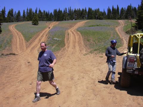 1991 Ford Bronco - Two men stand near off-road vehicles on a dirt track with rolling hills and trees in the background.