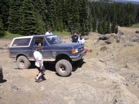 1991 Ford Bronco - A group of people are gathered around a lifted Ford Bronco navigating a rocky, off-road trail.