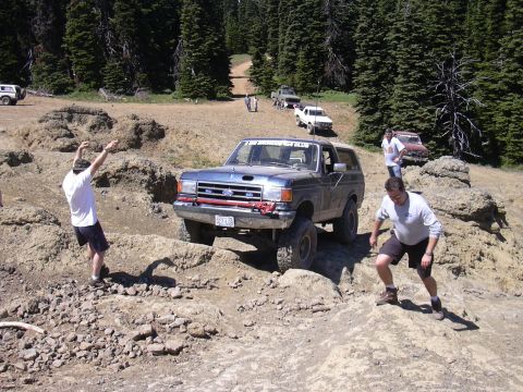 1991 Ford Bronco - A group of off-road vehicles, including a blue Ford Bronco, traverse a challenging dirt path in a wooded area.