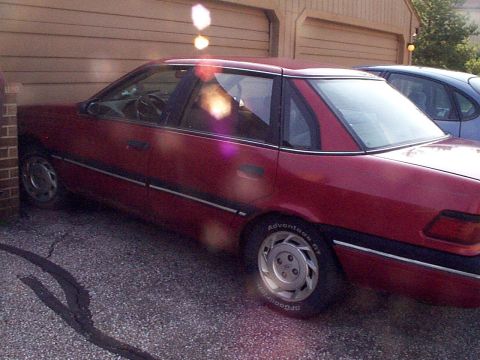 1999 Ford Taurus - This red sedan is parked in front of a garage. The body style suggests a 1999 model year, but it does not appear to be a Ford Taurus.