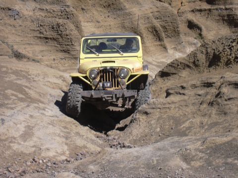 1991 Ford Bronco - A yellow Jeep CJ-7 navigates a challenging off-road trail in a rocky, arid landscape.