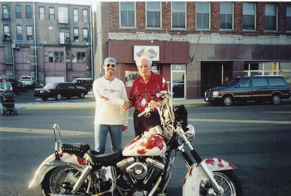Two men shake hands next to a custom motorcycle with a red and white paint job. A van is parked in the background