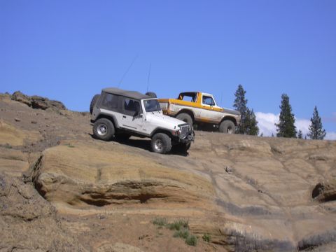 1991 Ford Bronco - Two off-road vehicles navigate a , rocky terrain under a clear blue sky.