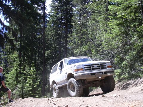 1991 Ford Bronco - A white Ford Bronco navigates a steep, rocky trail surrounded by trees.