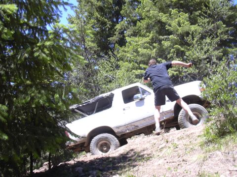 1991 Ford Bronco - A man stands by a white SUV as it navigates a steep, wooded incline.