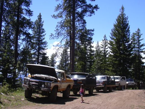 1991 Ford Bronco - A line of off-road vehicles, including a yellow and white pickup with its hood up, parked on a dirt road.