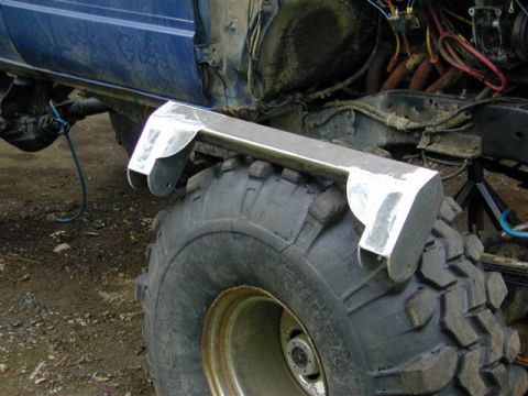 1985 Ford Bronco - A freshly fabricated bare-steel crossmember with welded end brackets rests on a large military-style mud tire on a white wheel, staged in f…