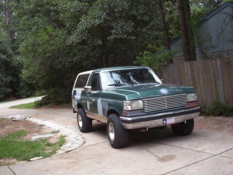 1989 Ford Bronco - This green SUV is parked in a driveway with trees and a wooden fence in the background. Body filler is visible on the front fenders and doo…