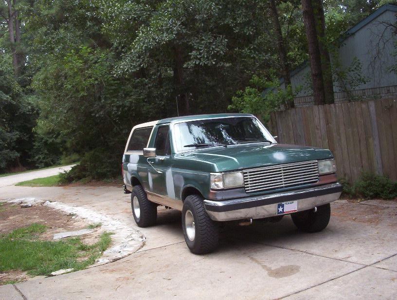 Green Ford Bronco with tan top and clear corner lenses parked on a driveway by a wood fence