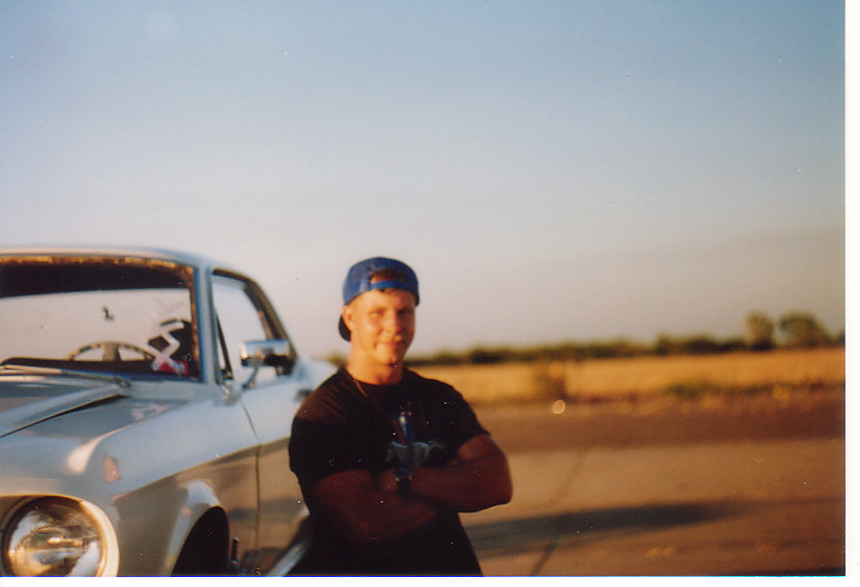 Guy in a blue baseball cap and black t-shirt leans against a silver classic car. Field and sky in background
