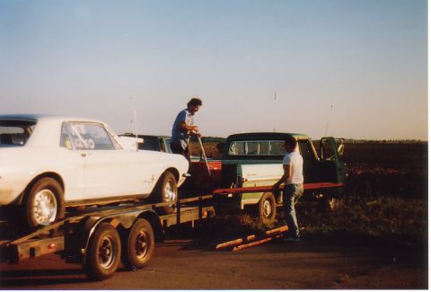 1968 Ford Mustang - Two men load a white 1965 Ford Mustang coupe onto a trailer towed by a green pickup truck under a clear sky.