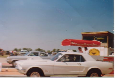 1968 Ford Mustang - A white 1968 Ford Mustang coupe is seen from the front-right, with another car partially visible behind it under a blue sky.