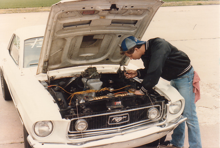 Man working on the V8 engine bay of a white classic Ford Mustang with hood up
