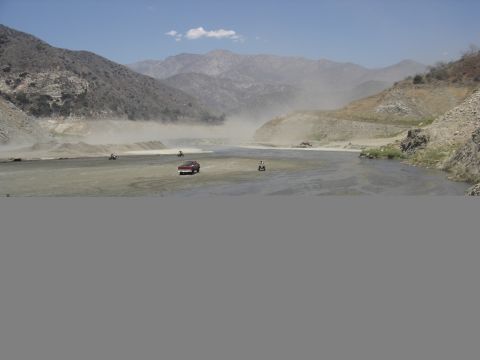 1990 Ford Bronco - Trucks and ATVs kick up dust crossing a wide dry riverbed in a rugged mountain canyon under a partly cloudy sky.