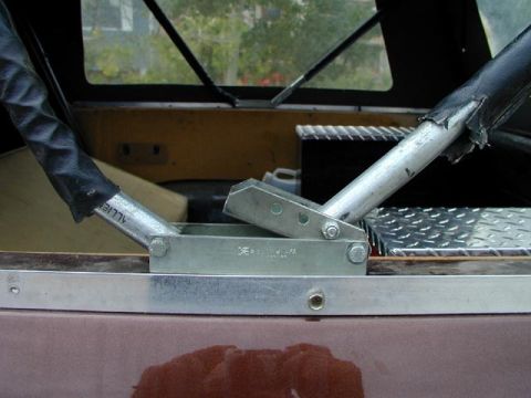 1979 Ford Bronco - Close-up of a galvanized soft-top hinge bracket bolted to the bed rail of a maroon truck, with two tubular bow arms pivoting on it and torn…
