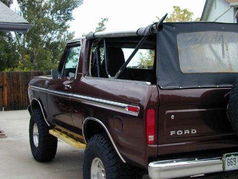 1979 Ford Bronco - The rear quarter of a dark maroon full-size Ford Bronco with 'FORD' lettering on the tailgate, a folded soft top over a roll bar, a chrome…