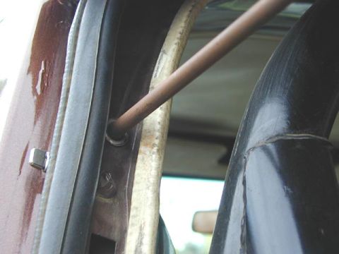 1979 Ford Bronco - Tight close-up inside a maroon vehicle's door opening showing a bare metal rod anchored to the B-pillar area, angling up past a weathered b…