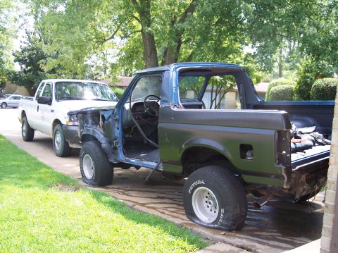 1990 Ford Bronco - A disassembled 1990 Ford Bronco sits with flat tires beside a white pickup truck in a driveway.