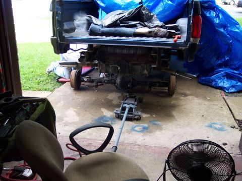 1990 Ford Bronco - Vehicle's rear axle and suspension are exposed and being worked on, with a jack supporting the frame and a blue tarp covering part of the v…