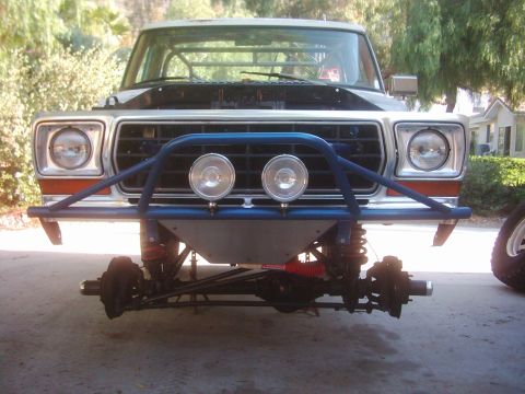 1978 Ford Bronco - Front view of a white late-1970s Ford 4x4 up on jack stands with both front wheels removed, exposing the solid front axle, coil springs, hu…