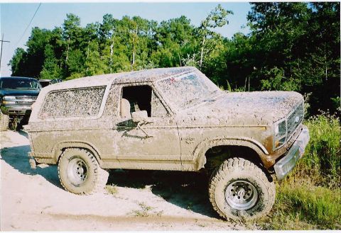1984 Ford Bronco - This heavily mud-caked 1984 Ford Bronco is seen off-road, with another SUV partially visible in the background.