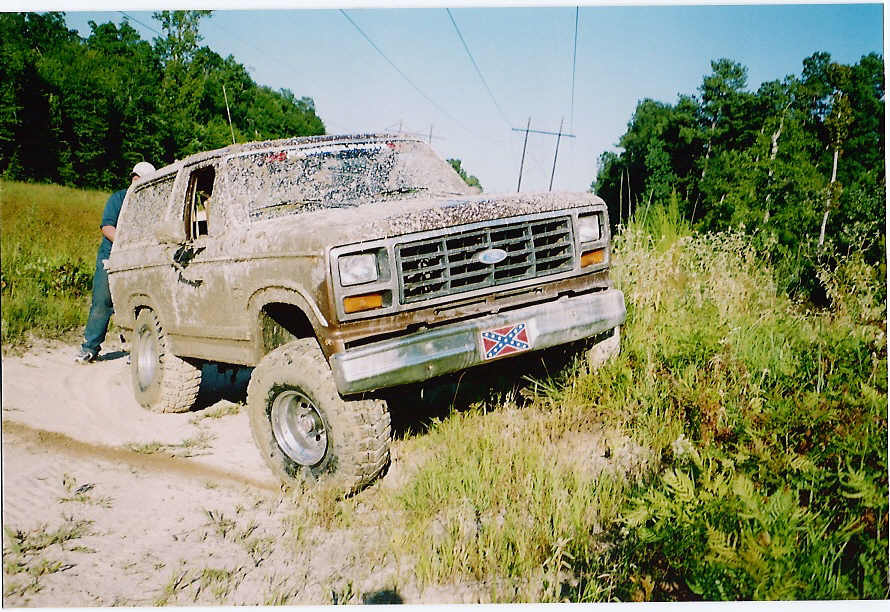 Mud-covered 1980s Ford Bronco with large tires on a dirt path, trees in background