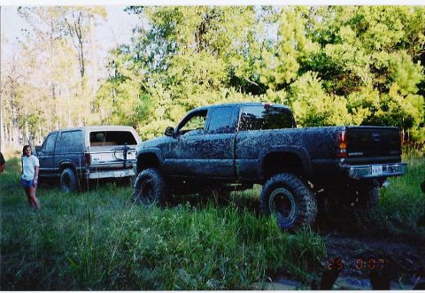 1984 Ford Bronco - Two off-road vehicles are covered in mud, parked in a grassy area with trees overhead.