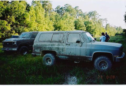 1984 Ford Bronco - Two SUVs are parked in a muddy, grassy area with trees in the background.