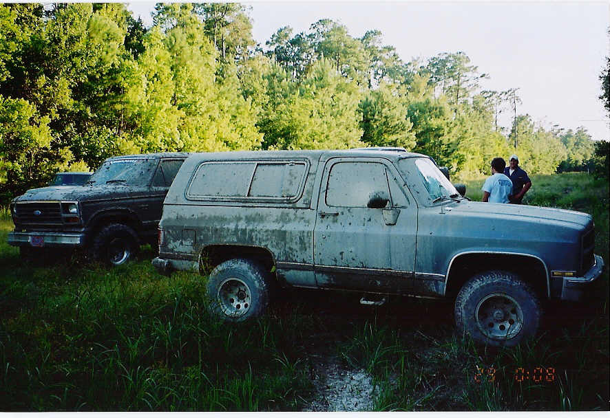 Two dirty SUVs parked in tall grass, one in foreground is blue-grey, the other behind it is dark brown