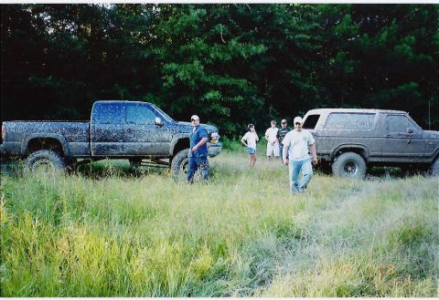 1984 Ford Bronco - Two muddy trucks, a Ford Bronco and a pickup, sit in a field with a group of people nearby.