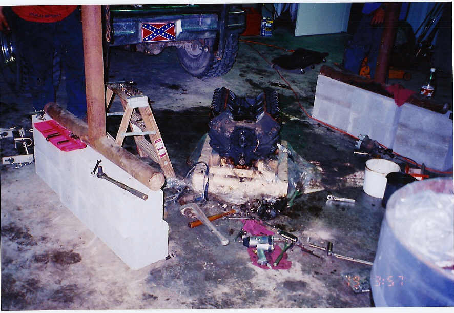 Engine block on floor, tools scattered around. Vehicle with Confederate flag sticker visible in background