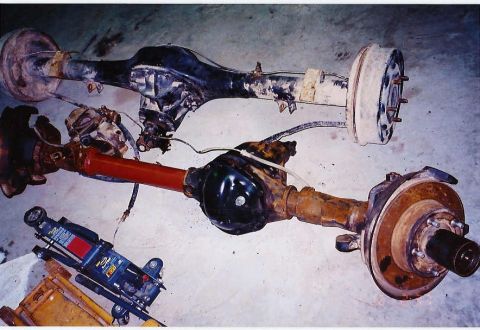 1984 Ford Bronco - Two vehicle axles are shown, one a red driveshaft, alongside a hydraulic jack.