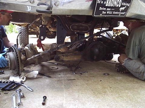 1989 Ford Bronco - Mechanics work on the rear axle of a 1989 Ford Bronco, confirmed by a license plate and visible vehicle components.