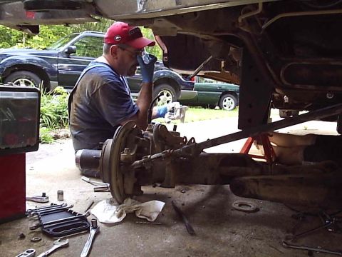 1989 Ford Bronco - Mechanic working on vehicle undercarriage with tools and parts laid out. Another vehicle is parked nearby.