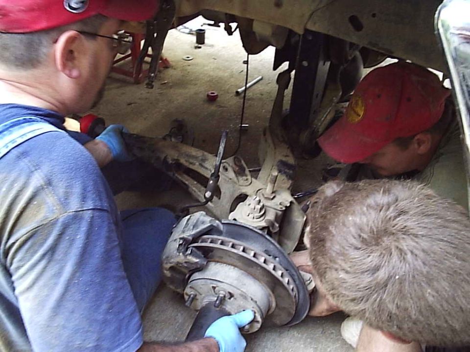 Three people working on a truck's front hub, rotor and steering knuckle from the ground