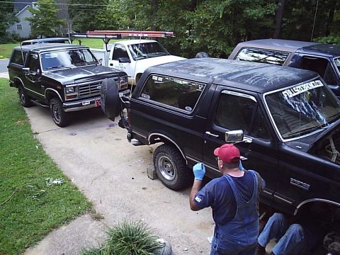 1989 Ford Bronco - A black 1989 Ford Bronco is parked in a driveway with a man working on its side, alongside other vehicles.