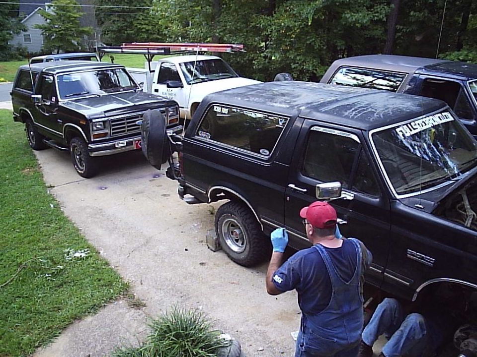 Three black Ford Broncos in a driveway with people working under one, front wheel removed
