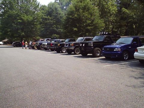 1989 Ford Bronco - A group of trucks and SUVs are parked in a lot on a sunny day, with trees in the background.