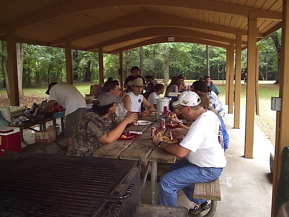 People eating at picnic tables under a covered pavilion, with trees in the background