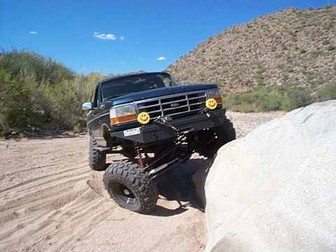 1994 Ford Bronco - This modified Ford truck navigates a rocky desert terrain with its front tire on a large boulder.