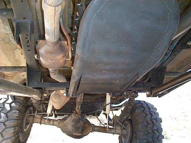 Underbody view: black fuel-tank skid plate, catalytic converter, and solid axle with mud-terrain tires