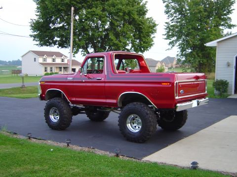 1978 Ford Bronco - Red Ford Bronco on a tall suspension lift with chrome wheels and mud tires, parked rear-quarter in a residential driveway.